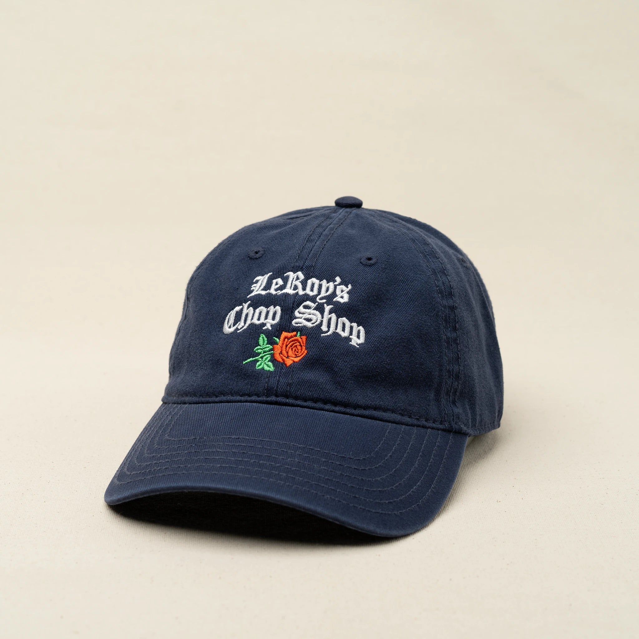 A navy blue baseball cap with an embroidered logo that reads 'LeRoy's Chop Shop' in white cursive, featuring a red rose design underneath.