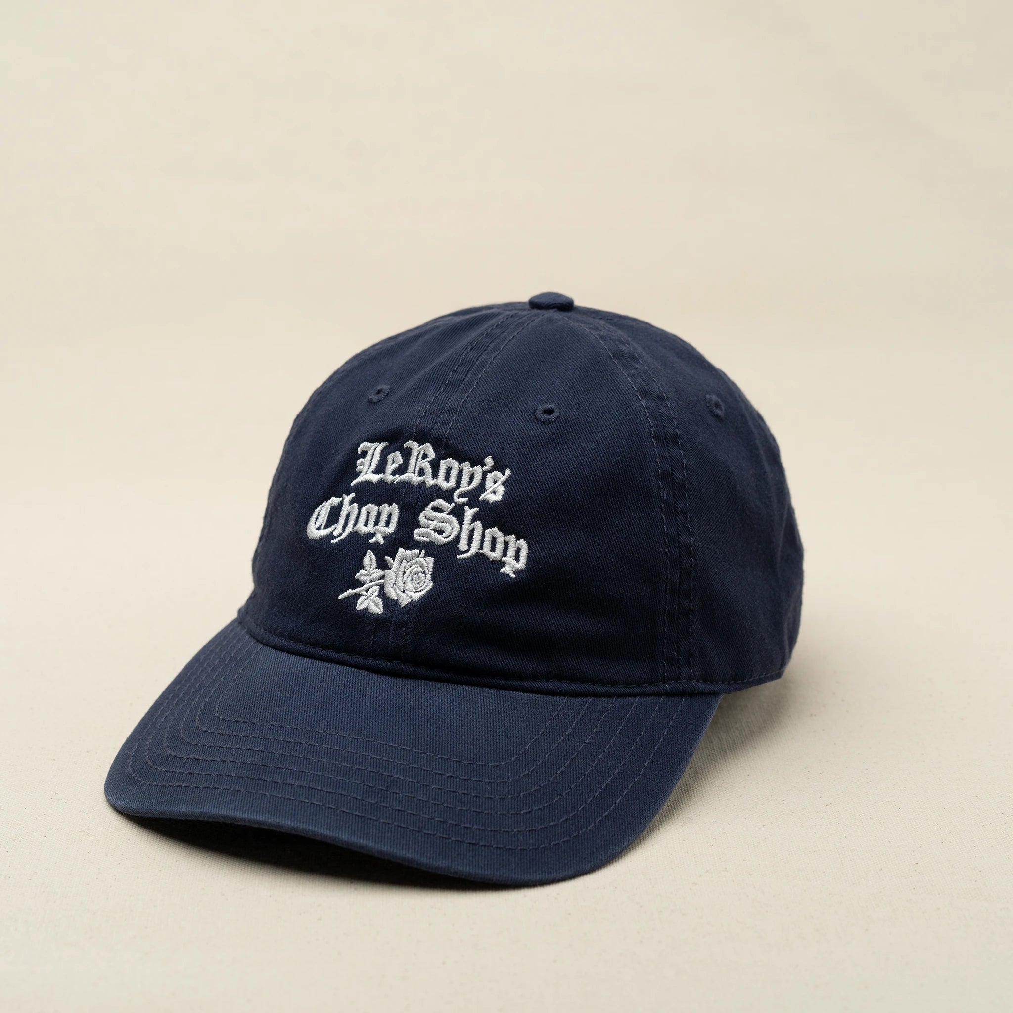 A navy blue baseball cap with an embroidered logo that reads 'LeRoy's Chop Shop' in white cursive, featuring a rose design underneath.
