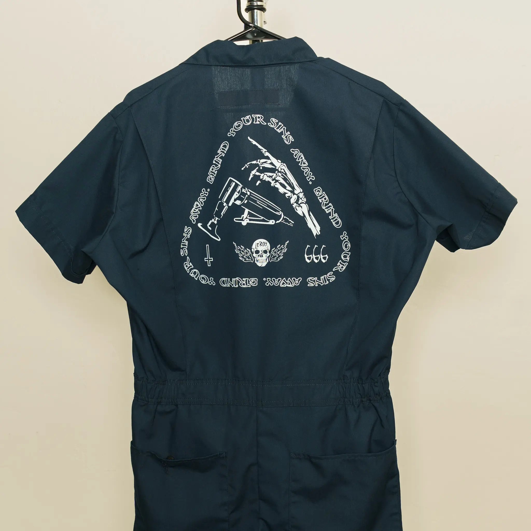 The back of a dark blue pair of coveralls hanging on a hanger, featuring a white graphic print with a skeleton hand holding a die grinder, the text 'GRIND YOUR SINS AWAY' arranged in a triangular pattern, '666', a skull with wings, and a cross.