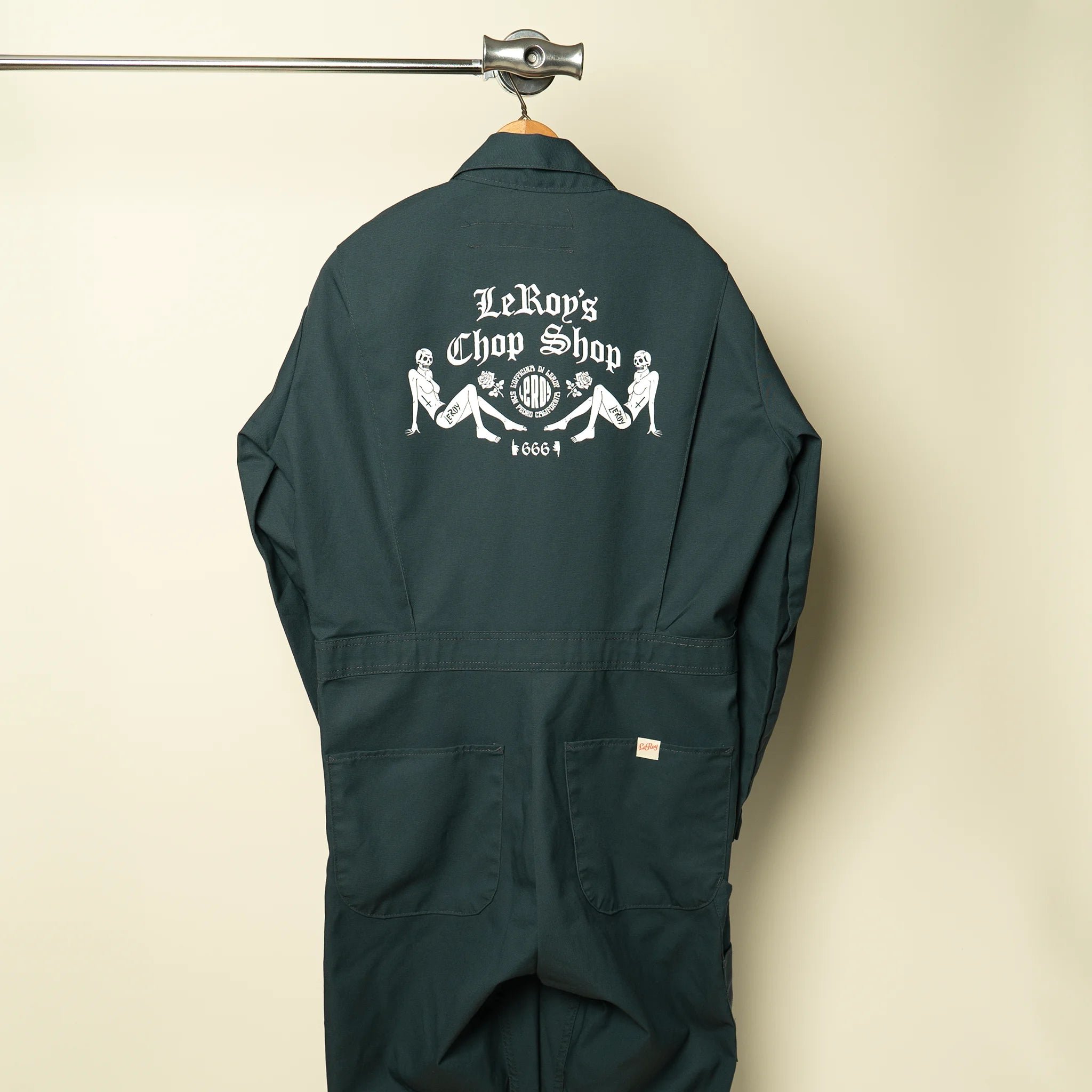 A pair of dark green coveralls hanging on a wooden hanger against a neutral background, featuring a large white graphic print on the back with 'LeRoy's Chop Shop' in gothic font, reclining figures, and the number '666'.