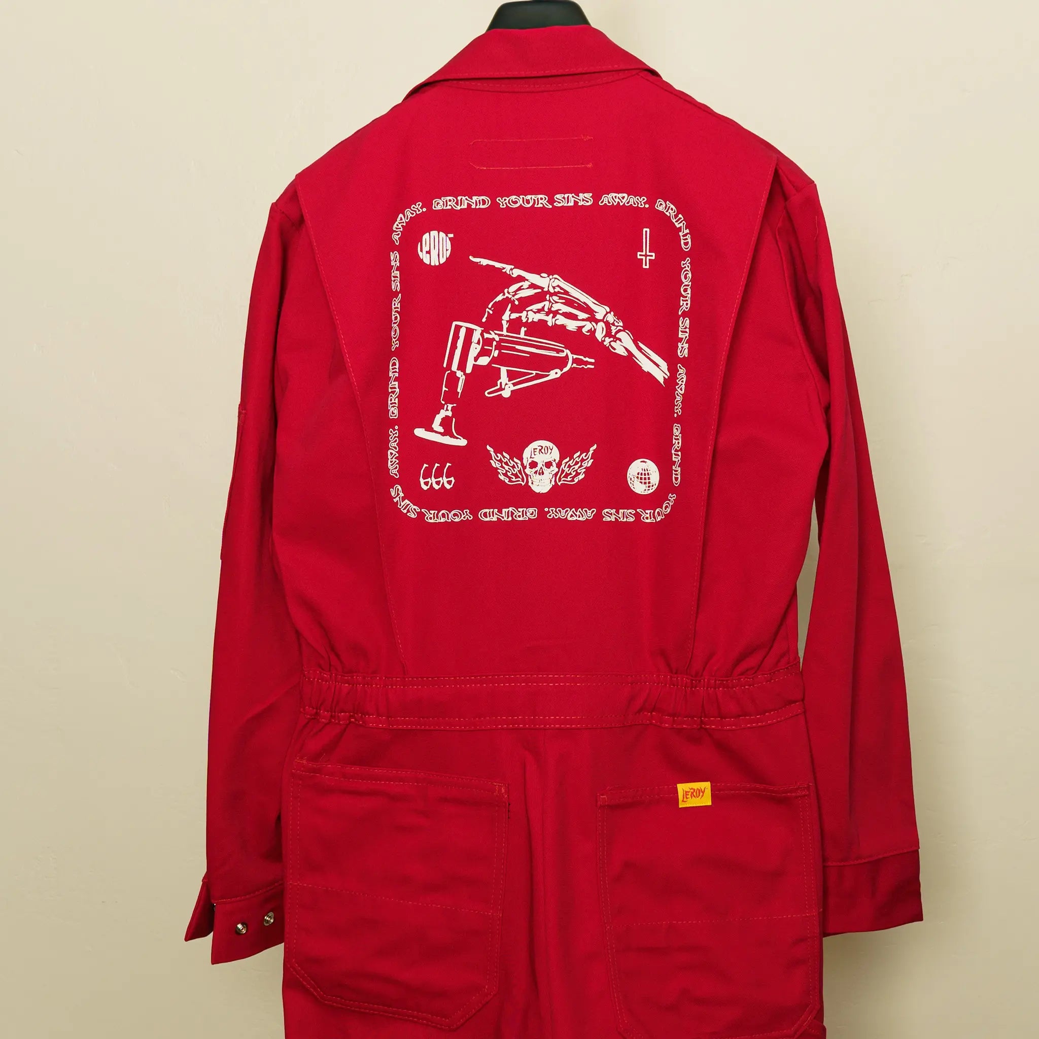 The back of red coveralls hanging on a hanger, featuring a large white graphic print with a skeleton hand holding a die grinder, the text 'GRIND YOUR SINS AWAY', '666', a skull with wings, and a cross.