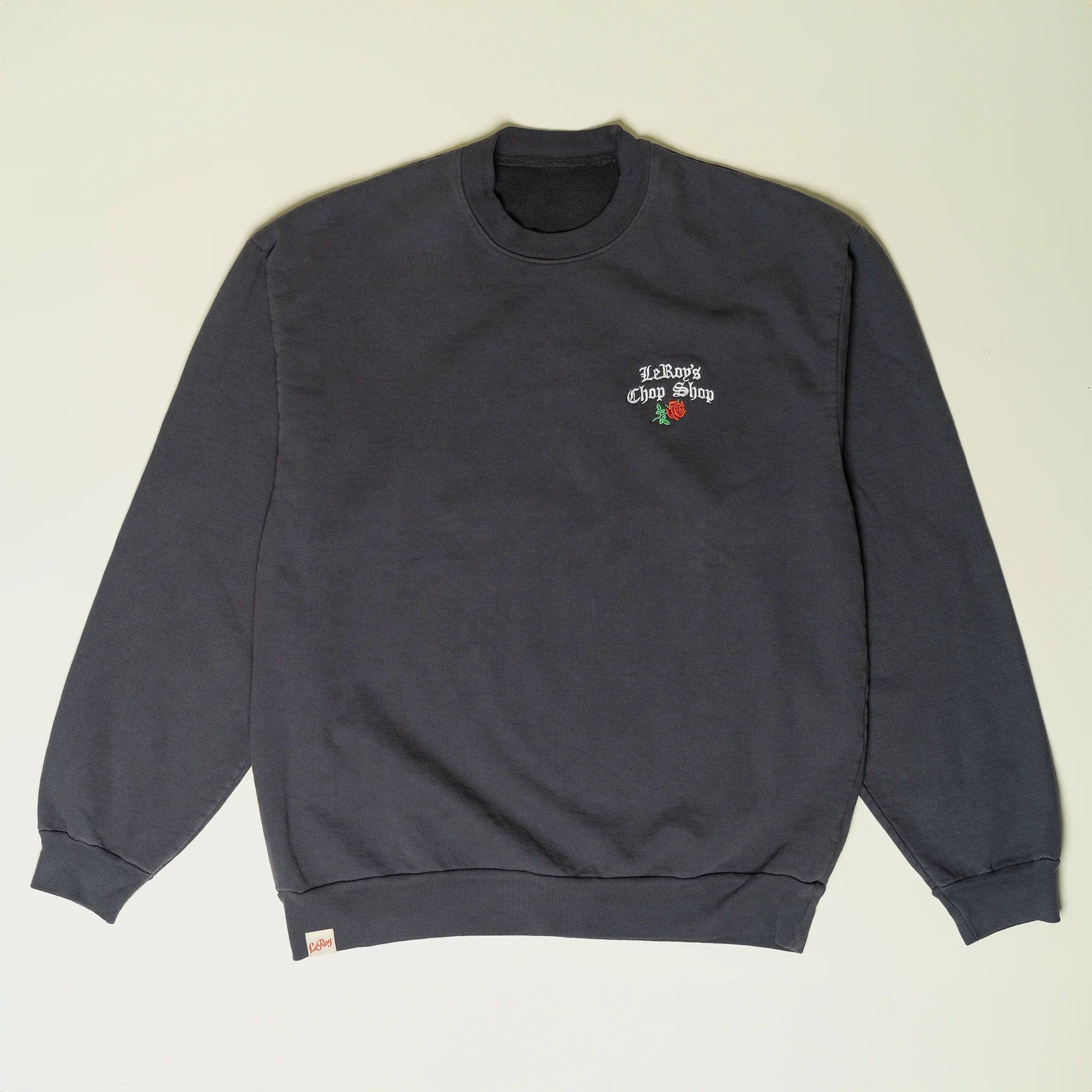A dark gray crewneck sweatshirt laid flat on a light background, featuring a small embroidered logo with 'LeRoy's Chop Shop' and a rose on the upper right chest, and a woven label on the lower left.