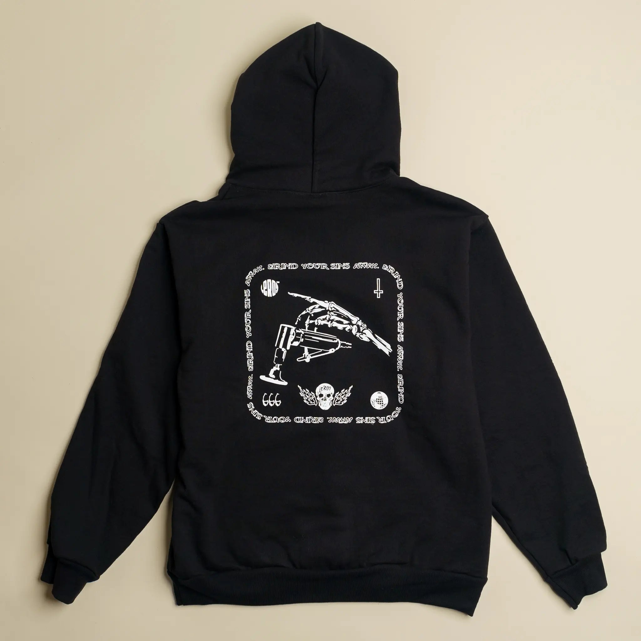 A black hoodie laid flat on a light background, featuring a white graphic print on the back with a skeleton hand holding a die grinder, the text 'GRIND YOUR SINS AWAY' arranged in a square pattern, '666', a skull with wings, and a cross.
