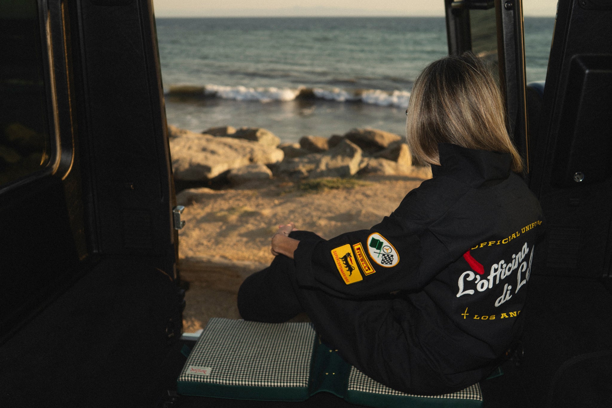 Person sitting on a leisure cushion inside a g-wagon looking out at a coastal landscape with waves and rocks.