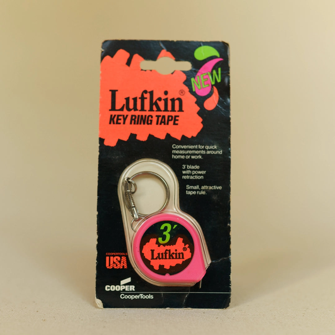 A vintage Lufkin Key Ring Tape measure package, featuring a pink tape measure with a silver ring, encased in a black and pink cardboard display with text describing its features and branding.