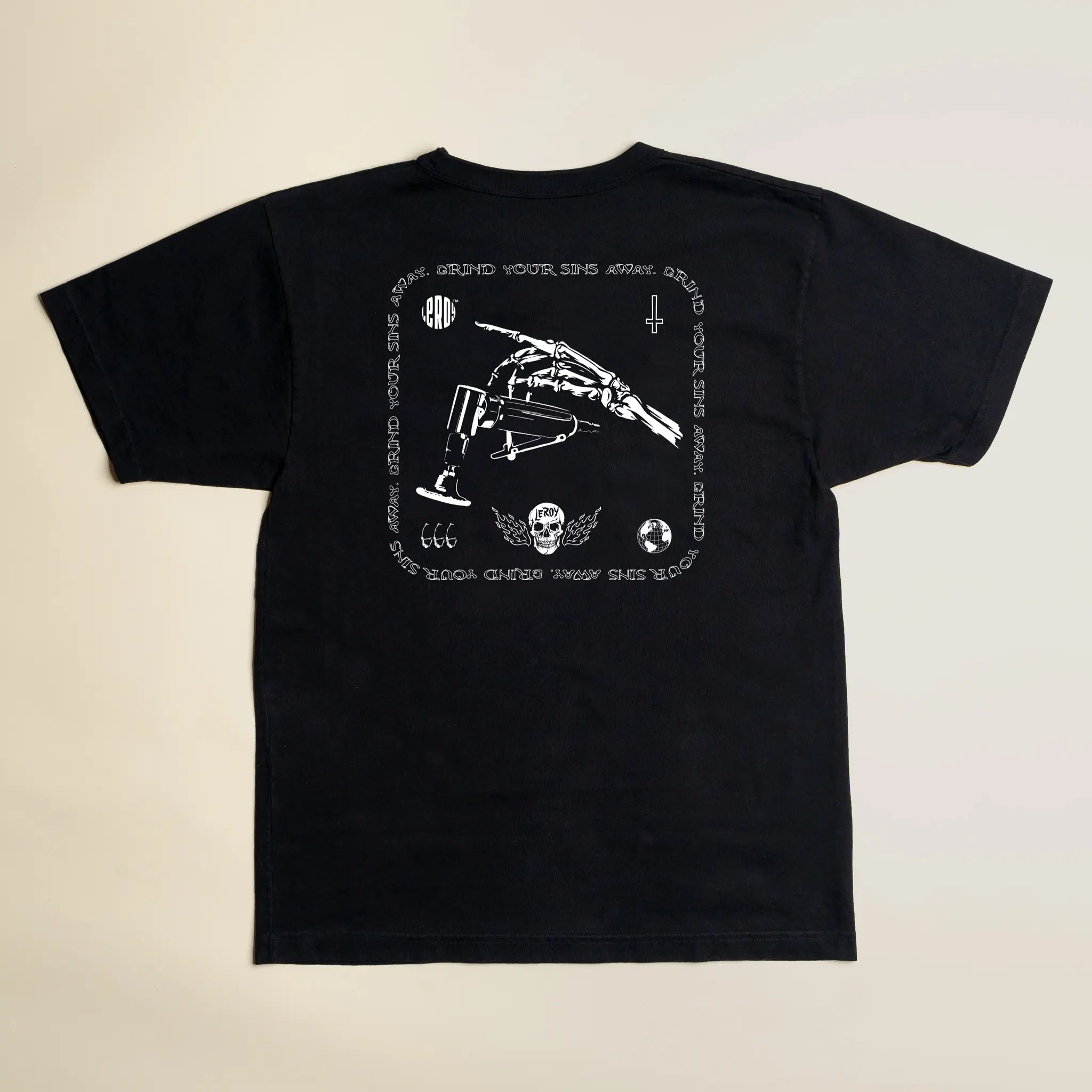 A black t-shirt laid flat on a light background, featuring a white graphic print on the back with a skeleton hand holding a die grinder, the text 'GRIND YOUR SINS AWAY' arranged in a square pattern, '666', a skull with wings, and a cross.