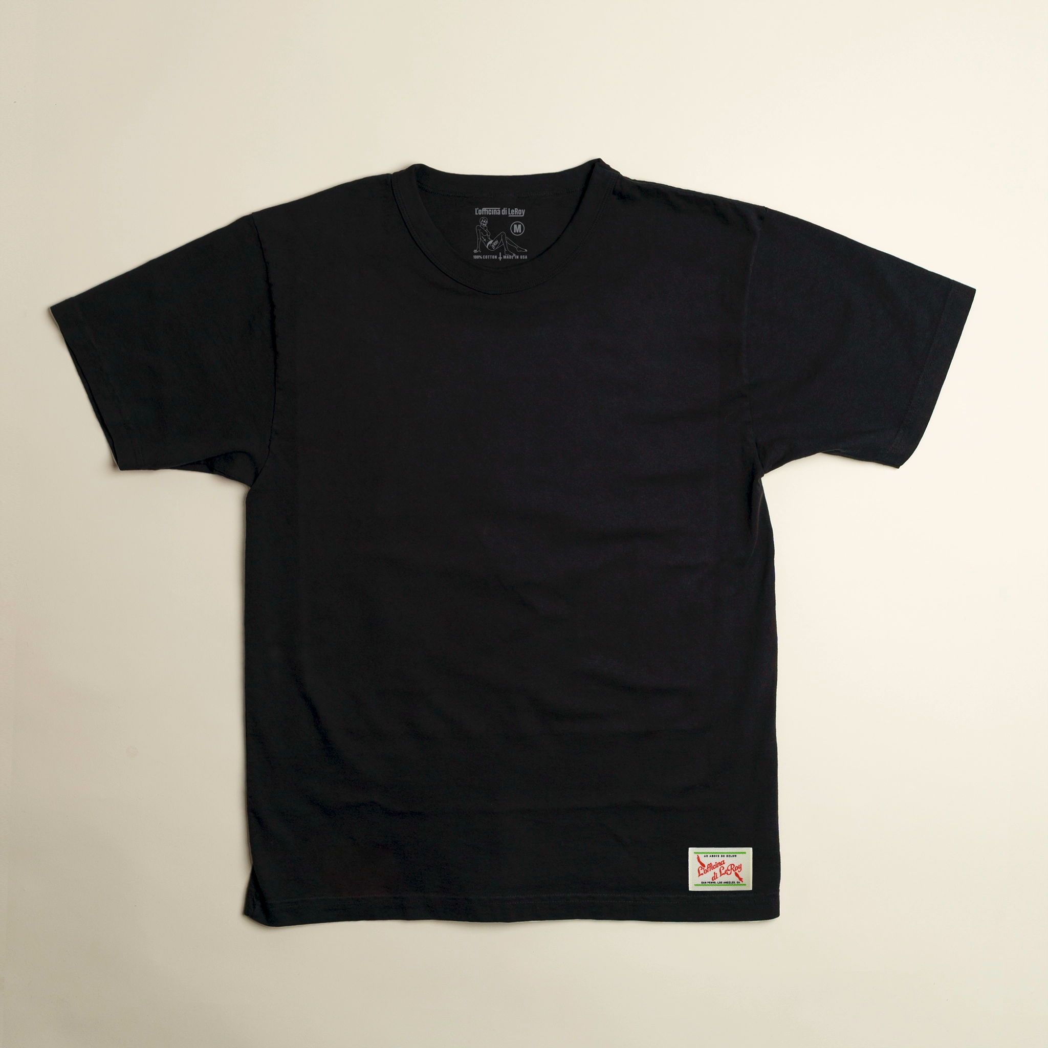 A black t-shirt laid flat on a light background, featuring a small woven label with red lettering on the lower right.